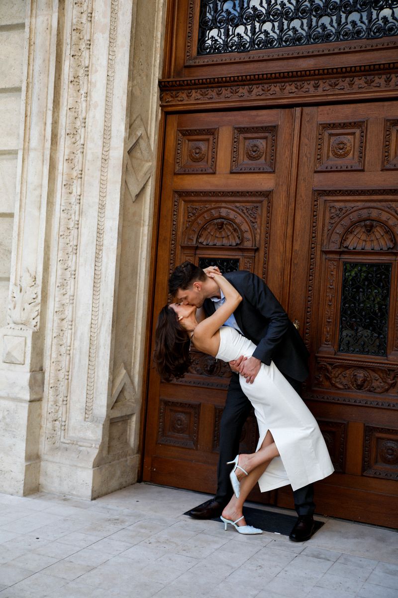 A couple kissing in front of a carved wooden door, she leans back in a white dress