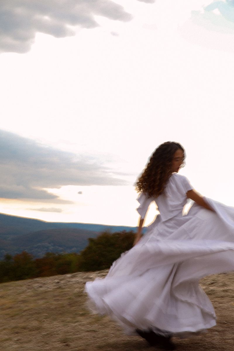 A woman in a white dress twirling on a hilltop at dusk