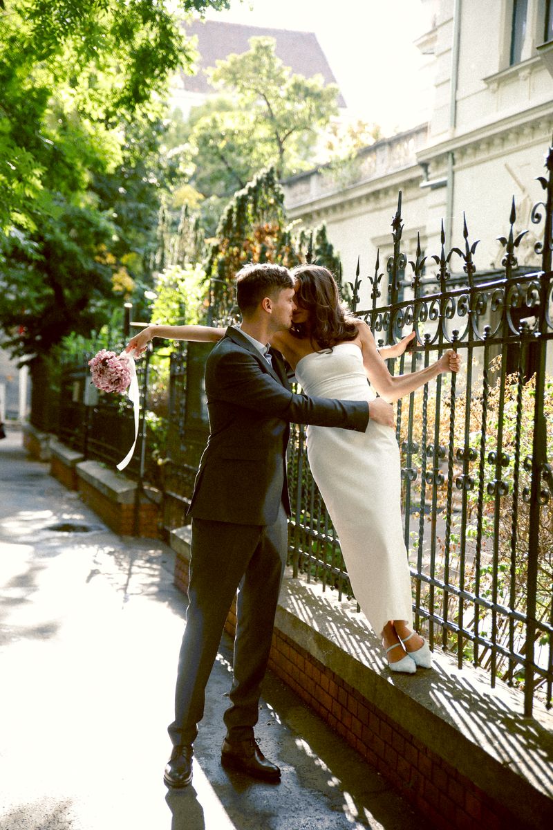 A couple kissing by a wrought-iron fence, she holds a bouquet out to the side