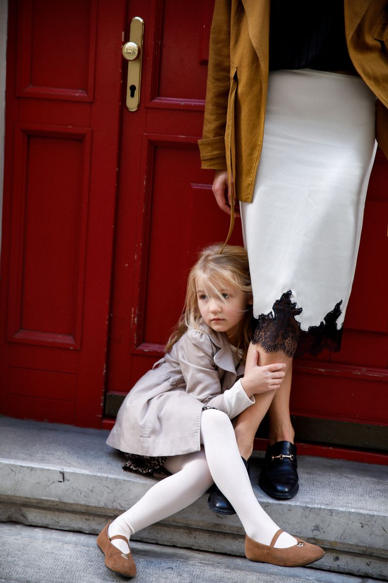 A girl in a trench coat on the step of a red door, her mother beside her