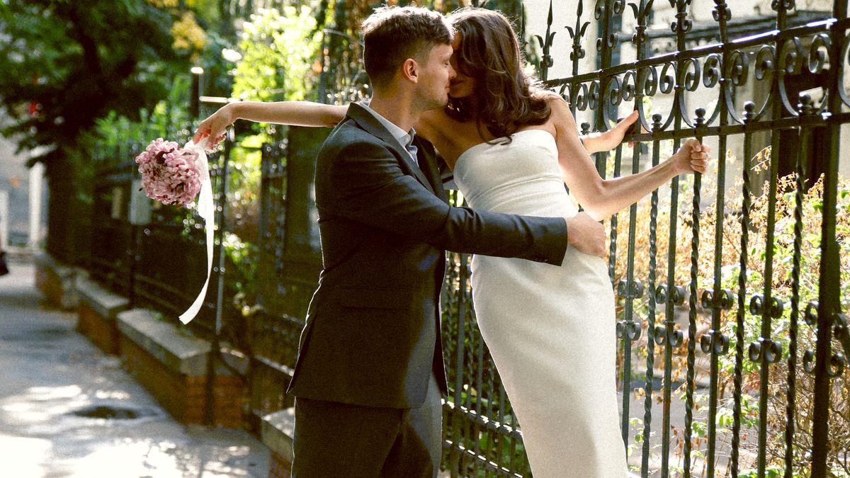 A couple kissing by a wrought-iron fence, she holds a bouquet out to the side